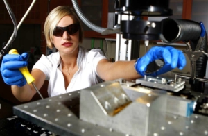 An image of a blonde woman wearing a white blouse and dark safety glasses working with a CNC milling machine