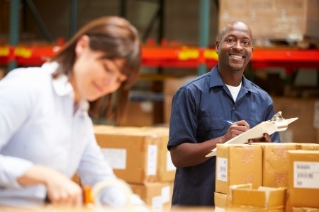 Two happy people, a black man and a caucasian woman marking and packaging contract manufacturer products in a warehouse. They are both smiling widely. 