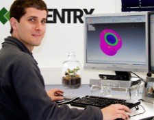 An image of a man sitting in front of his computer monitor, he is turned to look at the camera and is slightly smiling. On the monitor is a CAD part